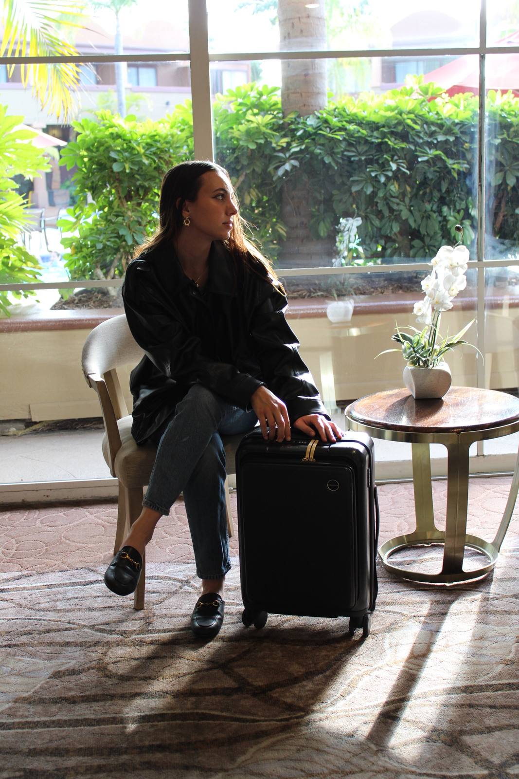 Model seated by a sunlit window with a black Champs carry-on