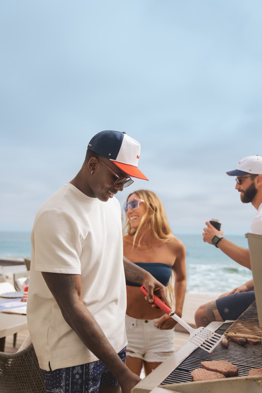Model in a Melin hat working the grill at a beachside cookout with friends