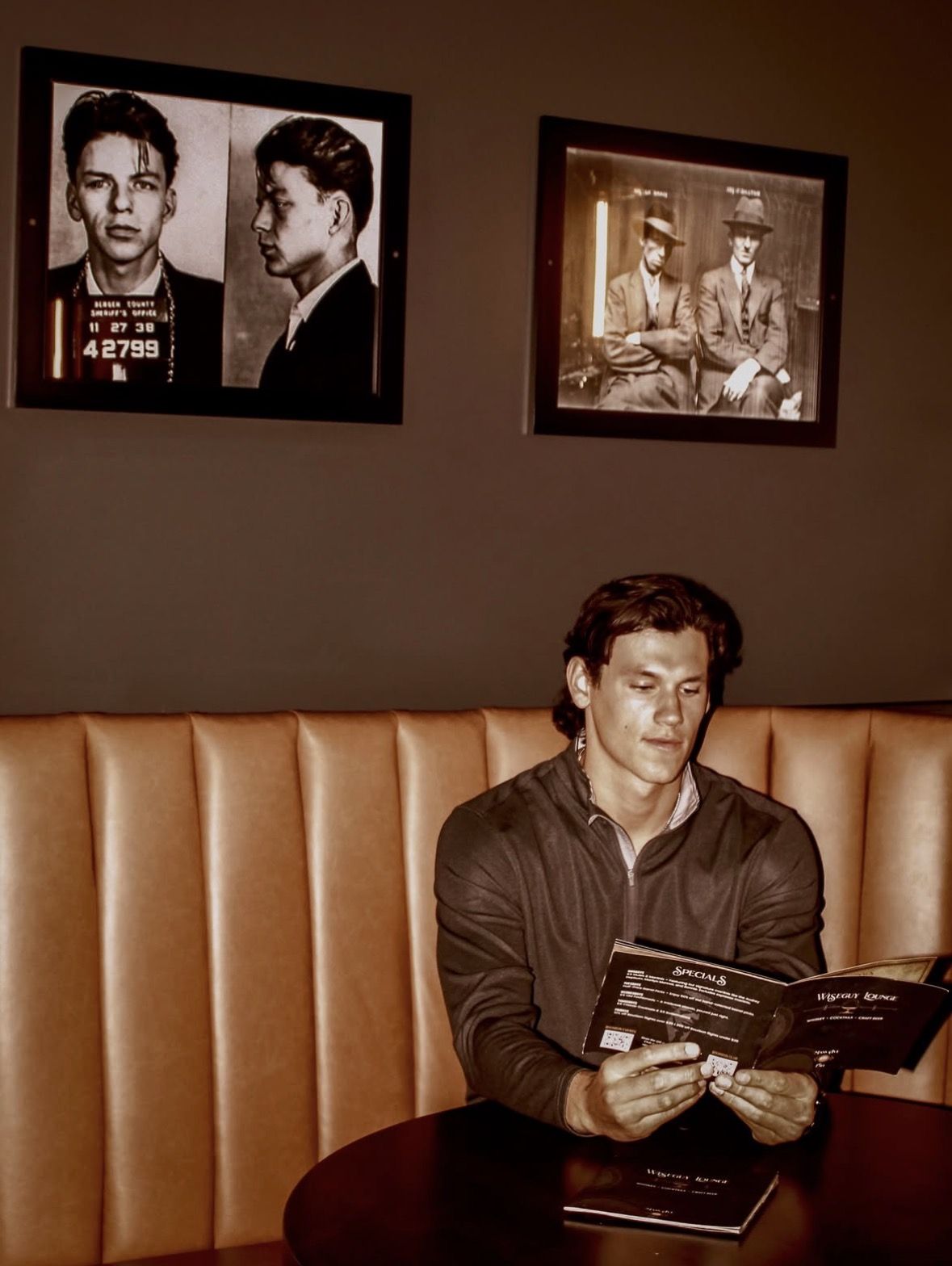 Guest reading the specials in a leather booth at Wiseguy Lounge, under framed vintage mugshots